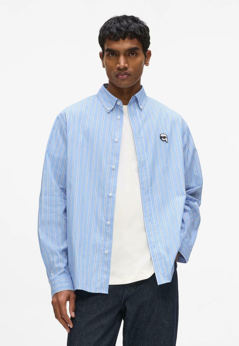 Homme portant une chemise à rayures verticales bleu clair sur un t-shirt blanc et un pantalon foncé, debout avec une main dans la poche.