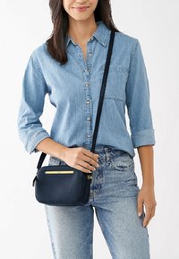 Vrouw in een lichtblauwe denim shirt en jeans, met een kleine zwarte crossbody tas met een gouden accent aan de voorkant.