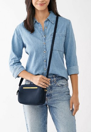 Žena v světle modré džínové košili a džínech, držící malou černou crossbody tašku s zlatým akcentem vpředu.