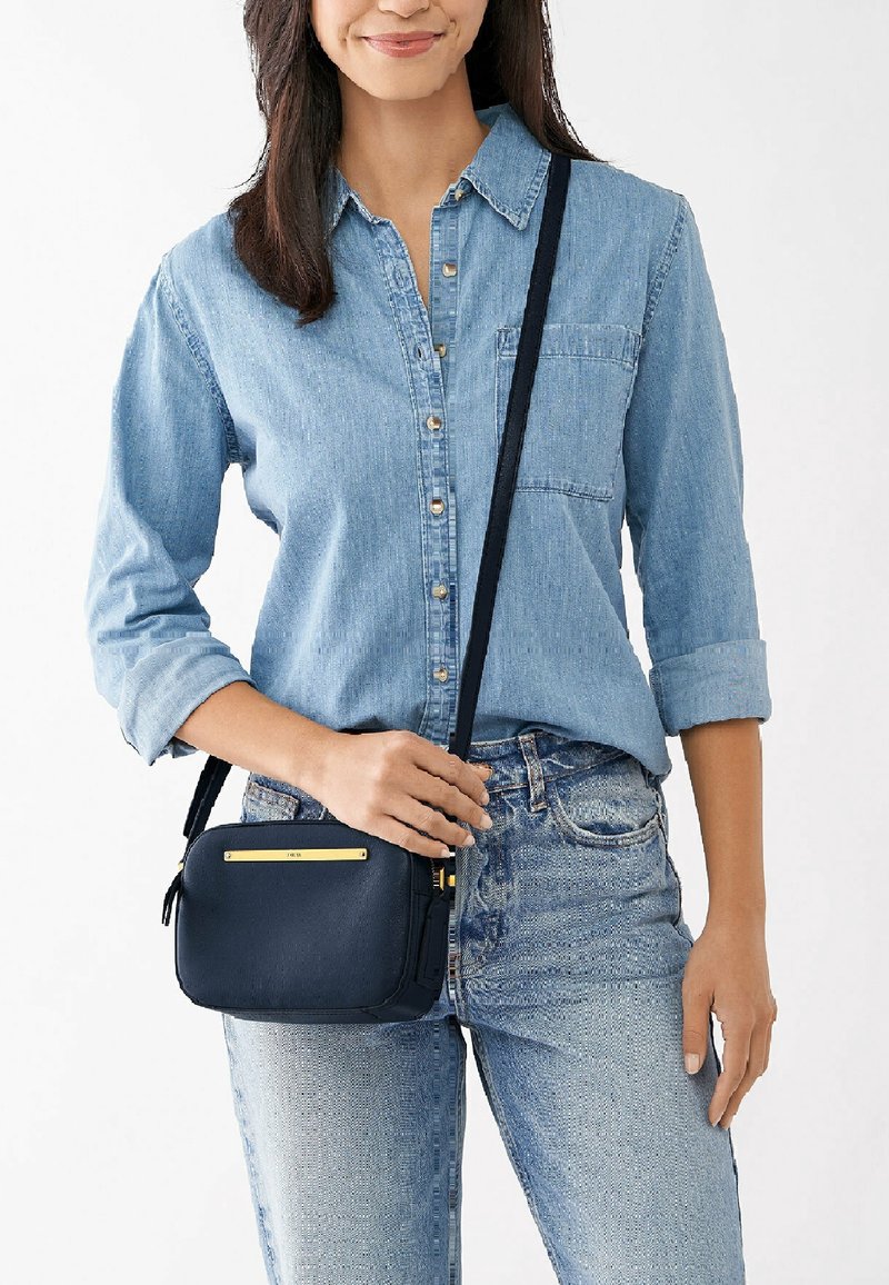 Vrouw in een lichtblauwe denim shirt en jeans, met een kleine zwarte crossbody tas met een gouden accent aan de voorkant.