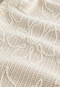 Tissu beige texturé avec motifs floraux en relief et un bouton blanc partiellement visible dans le coin supérieur gauche.