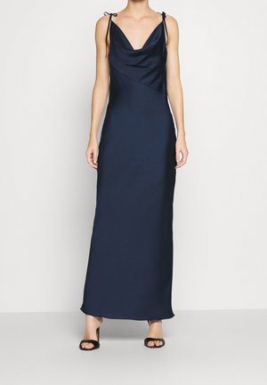 Femme portant une robe longue bleu marine sans manches avec de fines bretelles à nouer et des talons ouverts noirs.
