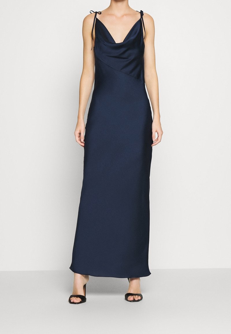Femme portant une robe longue bleu marine sans manches avec de fines bretelles à nouer et des talons ouverts noirs.
