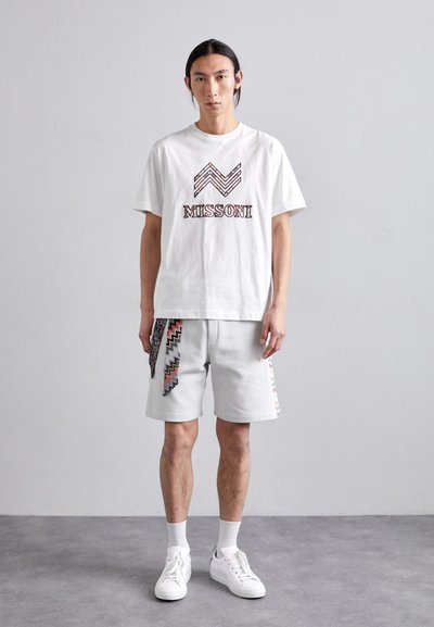 T-shirt en coton blanc avec le logo "MISSONI" et un motif en zigzag. Shorts gris avec des accents zigzag multicolores sur les côtés. Baskets blanches.