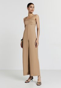 Frau steht in einem ärmellosen, sandfarbenen Jumpsuit mit weitem Bein und schwarzen Riemchenschuhen vor einem schlichten weißen Hintergrund.
