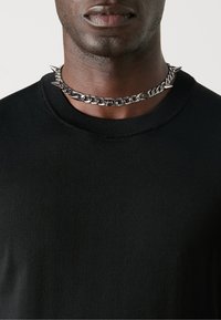 Jersey de cuello redondo negro hecho de tela texturizada, que presenta un collar de cadena plateada con picos y eslabones, colocado alrededor del cuello.