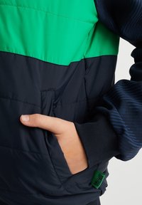 Vaatetus takki, jossa on väri-blokki muotoilu vihreänä ja laivastonsinisenä. Sisältää sivutaskun ja ribbeillä varustetut hihansuut, sekä pienen merkin helmassa.