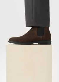 Brown suede Chelsea boot with elastic side panel, rounded toe, and black rubber sole, displayed on a light-colored pedestal.