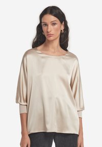 Beżowa satynowa bluzka z szerokim dekoltem, rękawami do połowy ramienia i luźnym krojem. Gładka faktura z delikatnym połyskiem. Noszona z ciemnymi dżinsami.