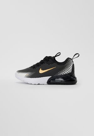 Zapatilla deportiva en blanco y negro con parte superior de malla, patrón de puntos, logo de Nike en dorado, suela acolchada y detalles de plástico negro.