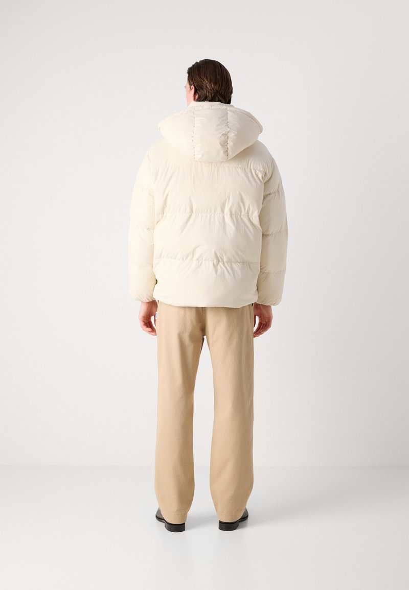 Giacca imbottita color crema con cappuccio, caratterizzata da sezioni trapuntate e una vestibilità comoda, abbinata a pantaloni beige e scarpe nere.