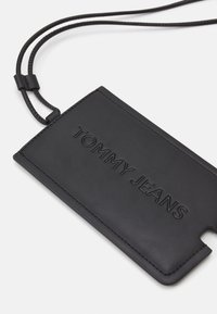Funda de teléfono de piel negra con el logo en relieve "TOMMY JEANS", que presenta una esquina recortada y una correa trenzada desmontable para facilitar el transporte.