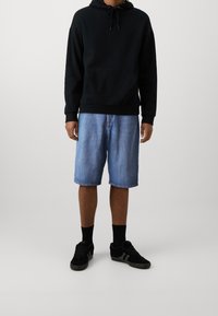 Sudadera negra con capucha y bolsillo frontal, combinada con shorts de denim azul claros. Zapatillas negras con calcetines oscuros completan el conjunto.
