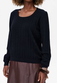 Czarny sweter prążkowany z długimi rękawami, okrągłym dekoltem i teksturowanym materiałem; noszony z brązową skórzaną spódnicą.