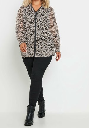 Femme portant un chemisier transparent imprimé léopard avec bordure noire, pantalon skinny noir et bottines noires, debout sur fond blanc.