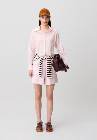 Joven con camiseta rosa claro y pantalones cortos, suéter a rayas atado a la cintura, zapatos de cuero marrón, gorra marrón, sosteniendo un bolso marrón oscuro.