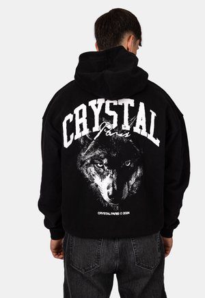 Osoba nosząca czarną bluzę z kapturem z dużym białym, pogrubionym napisem "CRYSTAL Paris" i szczegółową grafiką twarzy wilka w odcieniach szarości na plecach.