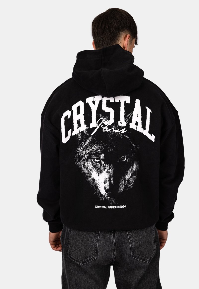 Personne portant un sweat à capuche noir avec un grand texte blanc en gras "CRYSTAL Paris" et un graphique détaillé en niveaux de gris représentant un visage de loup dans le dos.