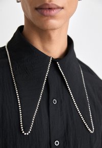 Camisa preta texturizada com um grande colarinho, apresentando um detalhe de corrente metálica adornada com pequenas pedras transparentes ao longo do decote.