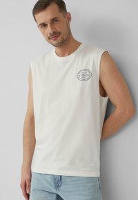 T-shirt sans manches en coton blanc avec un logo graphique rond sur la poitrine gauche. Le tissu est lisse et le design est simple sans accents supplémentaires.