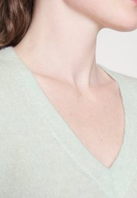 Gros plan sur le cou et le haut de la poitrine d'une personne portant un pull vert clair à col en V, avec la clavicule visible et une peau claire.