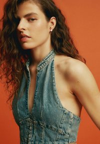 Mujer con cabello largo y rizado, vestida con una blusa de mezclilla azul sin mangas y pendiente de perlas, posando frente a un fondo naranja.