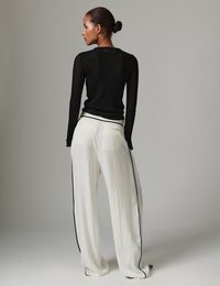 Γυναίκα με τα μαλλιά της σε κότσο, που φοράει μαύρο διάφανο ριμπ πλεκτό πουλόβερ και άσπρο παντελόνι με φαρδιά μπατζάκια και μαύρες πλαϊνές ρίγες, στέκεται μπροστά σε απλό γκρι φόντο.