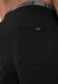 Svarta sweatshirtbyxor med ett grått elastiskt midjeband som har "STHUGE"-logotypen. Funktioner inkluderar en stor bakficka med förstärkt sömnad.