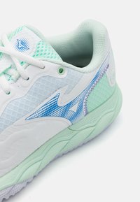 Baltās un piparmētru zaļas sporta kurpes ar sietveida augšdaļu, teksturētām akcentiem un zilu grafisko dizainu sānos. Baltās lentes un mīkstināta apmale.