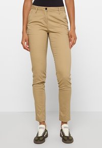 Personne portant un pantalon beige slim associé à des baskets Adidas noires et blanches, debout devant un fond clair uni.