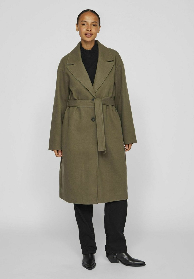 Olīvu zaļš garais mētelis ar pārmērīgiem atlokiem, jostasvietu, vienu pogu aizdari un plašām piedurknēm; apvienots ar melnu apģērbu un norādītām zābakiem.