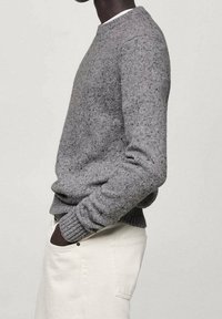 Personne portant un pull en maille grise texturée et un pantalon blanc, debout de profil avec les mains dans les poches, sur un fond uni.