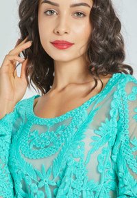Mujer con cabello rizado oscuro que lleva una blusa de encaje turquesa brillante, tocándose ligeramente el cabello, con un fondo neutro.