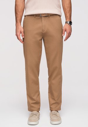 Homme portant un pantalon chino beige avec une ceinture noire, des chaussures beiges, un pull léger en maille et une montre argentée au poignet gauche.