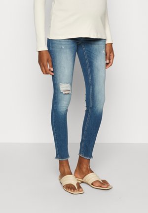 Jean slim bleu avec des patches usés sur les cuisses, associé à un haut à manches longues crème et des sandales ouvertes beige.
