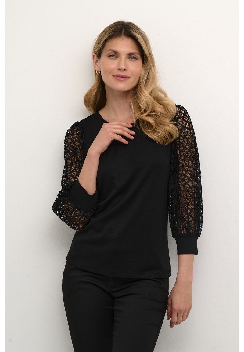 Haut noir avec de longues manches en dentelle présentant des motifs complexes, terminé par des poignets ajustés. Corps lisse et coupe décontractée.
