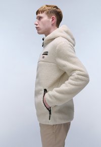 Cream-colored fleece jacket with a hood, featuring side pockets, a horizontal chest seam, and logo patch accents.