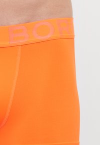 Oranžne bokserice, izdelane iz gladkega teksturiranega materiala, z širokim pasom, na katerem je v svetlejši oranžni barvi prikazano ime blagovne znamke "BORG".