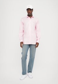 Απαλή ροζ, πουκάμισο με κουμπιά από βαμβακερό μείγμα, συνδυασμένο με ανοιχτό μπλε τζιν και λευκά αθλητικά παπούτσια. Ένα λευκό καπέλο διαθέτει ένα μικρό λογότυπο.