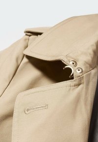 Gros plan du col d'un trench-coat beige avec des surpiqûres détaillées, des œillets métalliques et un crochet pour la fermeture.