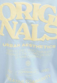 Světle modré tričko s velkým krémovým textem "ORIGINALS URBAN AESTHETICS", doplněné o menší text a ikonku zeměkoule.