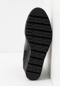 Semelle de chaussure noire avec une surface en caoutchouc texturée, présentant des rainures profondes et un talon plat. Le côté montre un accent en matériau à motif.