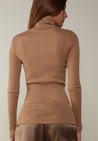 Haut à col roulé en maille côtelée, à manches longues, de couleur brun clair, présentant une coupe ajustée et une texture douce, associé à un tissu satiné en dessous.