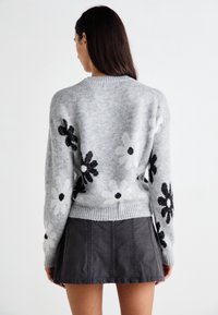 Femme aux longs cheveux foncés portant un pull gris avec des motifs floraux noirs et blancs et une jupe noire, de dos sur fond blanc.