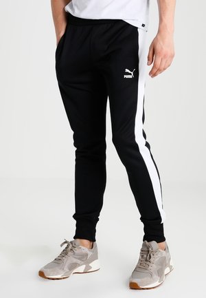 Homme portant un pantalon de survêtement noir Puma avec des bandes blanches sur les côtés et des baskets grises, mains dans les poches, debout sur un fond blanc uni.