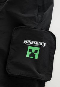 Fekete rövidnadrág nagyméretű zsebbel, a "Minecraft" logójával és egy zöld Creeper arc grafikával. Sima textúra, hétköznapi dizájn.