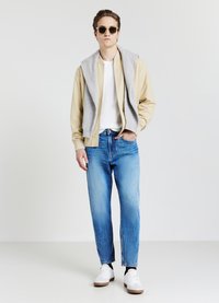 Giovane uomo che indossa occhiali da sole, giacca beige con un maglione grigio sulle spalle, t-shirt bianca, jeans blu e sneakers bianche, in piedi davanti a uno sfondo neutro.