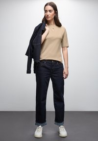 Beige kortærmet top med en nøgleruhul detalje, mørkeblå denim jeans med opkrøllede cuffs og hvide sneakers, ledsaget af en denimjakke.