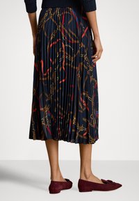 Nő sötétkék, pliszírozott midi szoknyában, melyen piros és barna láncminták láthatók, valamint bordó színű, masnival díszített szarvasbőr balerinacipőben, világos padlón áll.