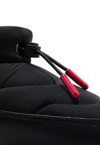 Zapatilla negra acolchada con forma redondeada, que cuenta con un cierre tipo toggle con cordón negro y puntas rojas, y una textura similar al cuero suave.